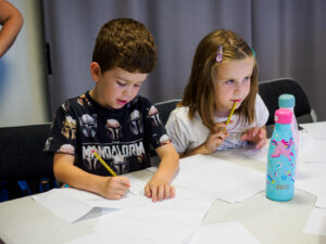 2 children in an arts and crafts workshop sat at the table with paper and pens in front of them.